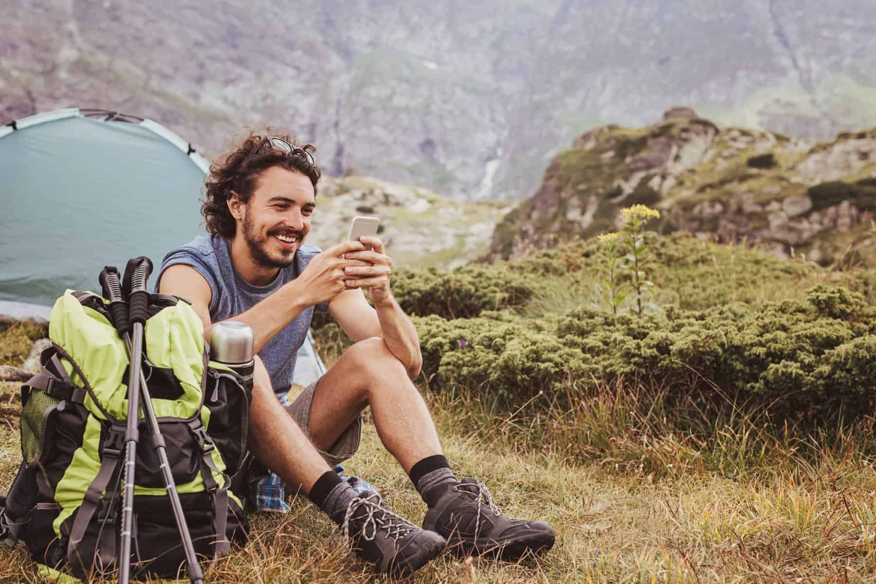 A man sitting in a field with a backpack and a smartphone, using the Campground App provided by Togo Group - one of the leading companies in creating innovative digital travel products.