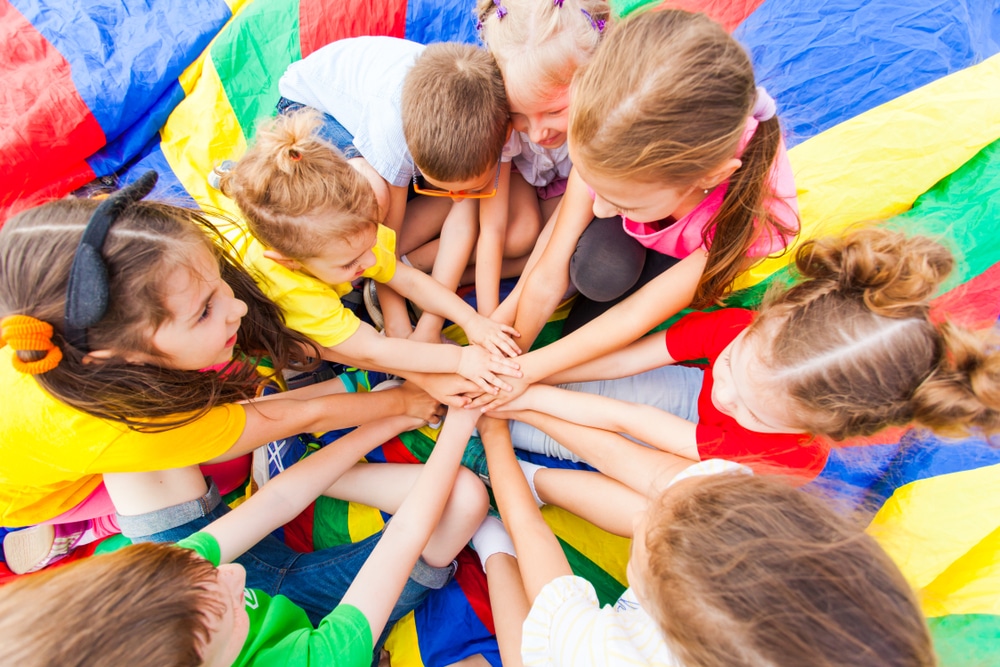 A group of children, including some kids with cancer, holding hands on a colorful parachute at a campground managed by a management company.