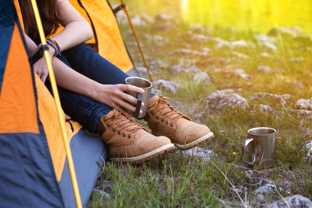 A woman, sporting Oboz shoes, is sitting in front of a tent with a cup of coffee.