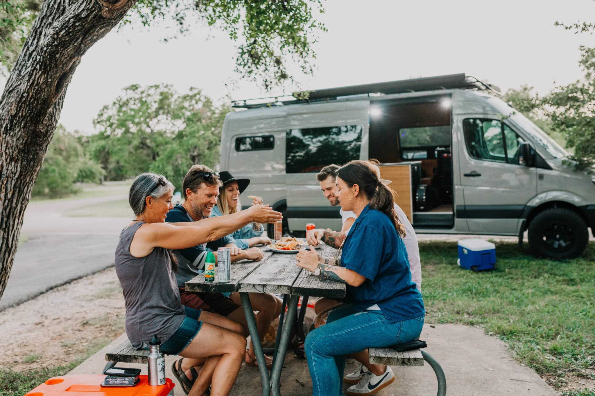 A group of people sitting at an outdoorsy picnic table near an RV.