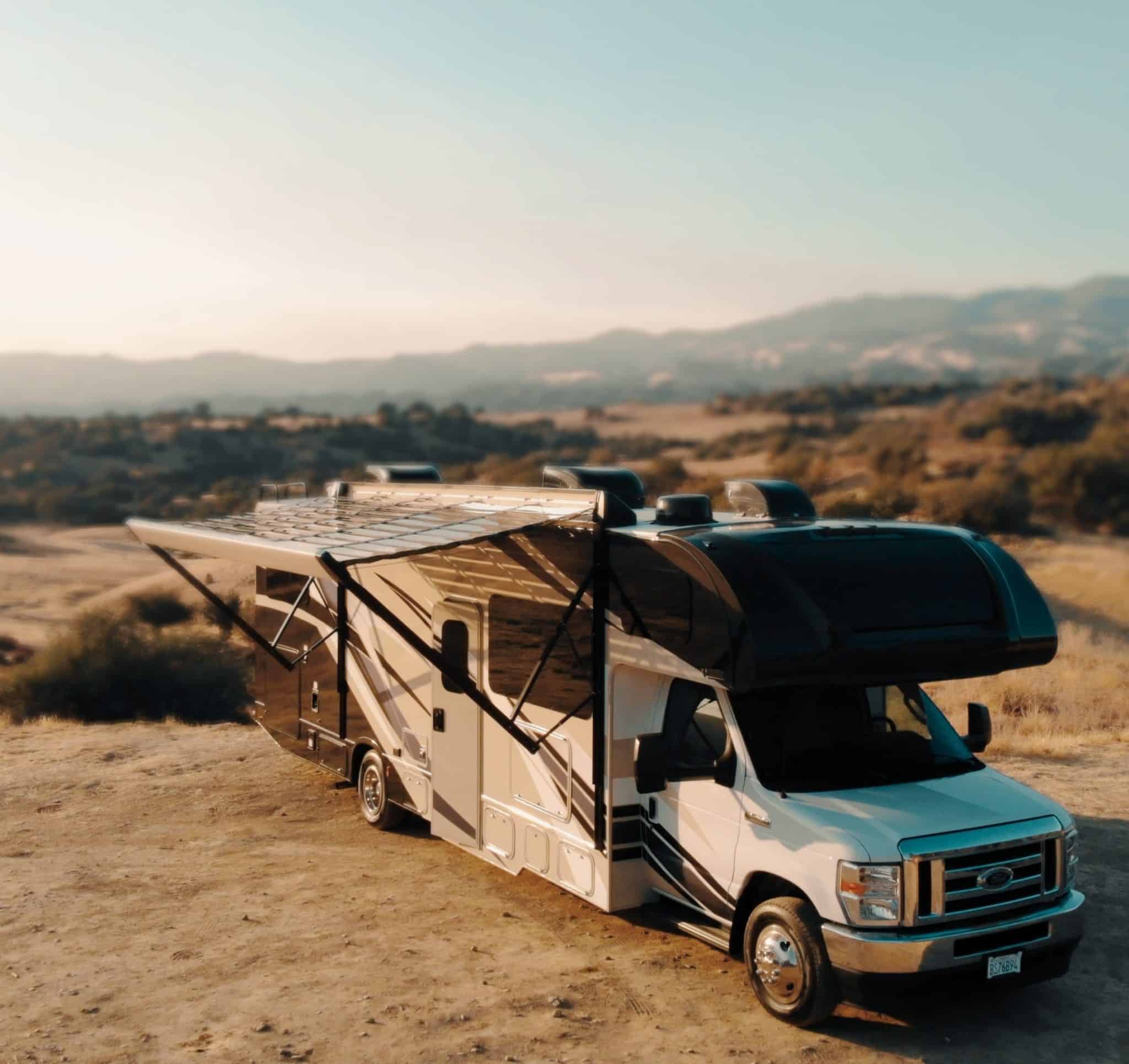 An RV equipped with a power-generating solar awning parked on a dirt road with mountains in the background.