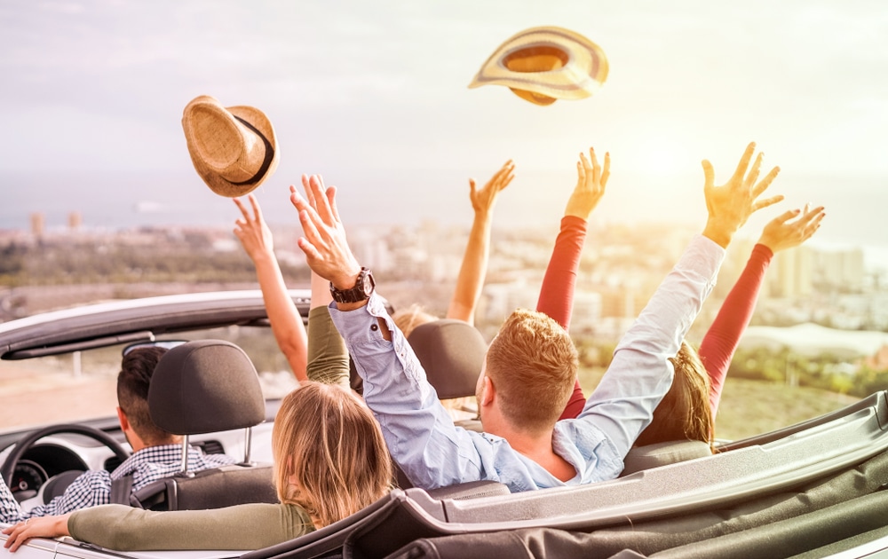 A group of people enjoying a joyride in a convertible car with their hands in the air, experiencing the thrilling AAA.
