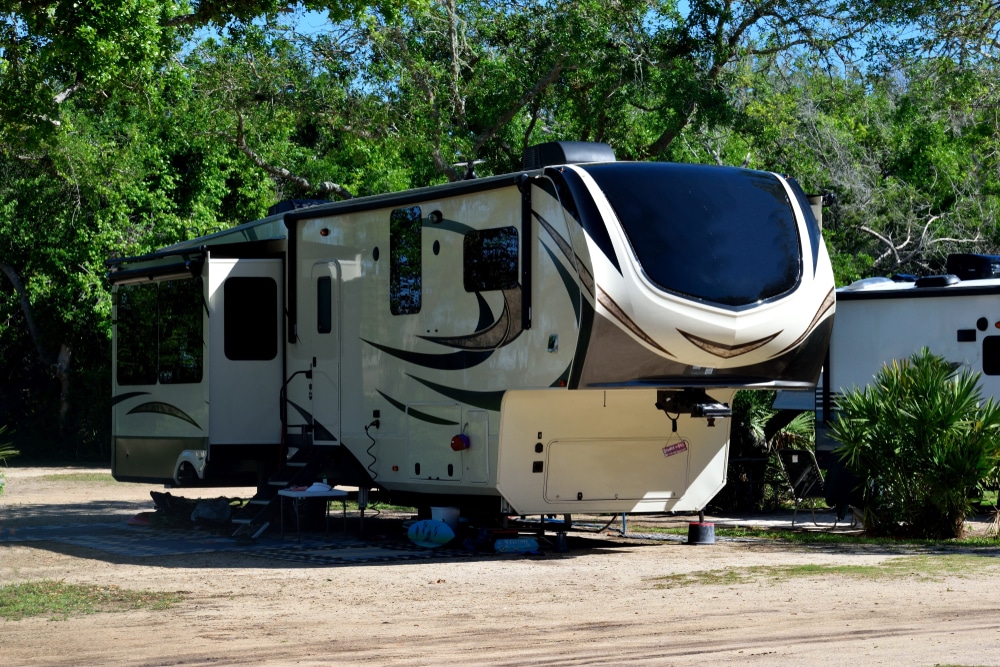 A luxury Fifth-Wheel RV parked in a dirt lot next to trees.