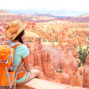 A woman with an orange backpack sits on a cliff overlooking bryce canyon.
