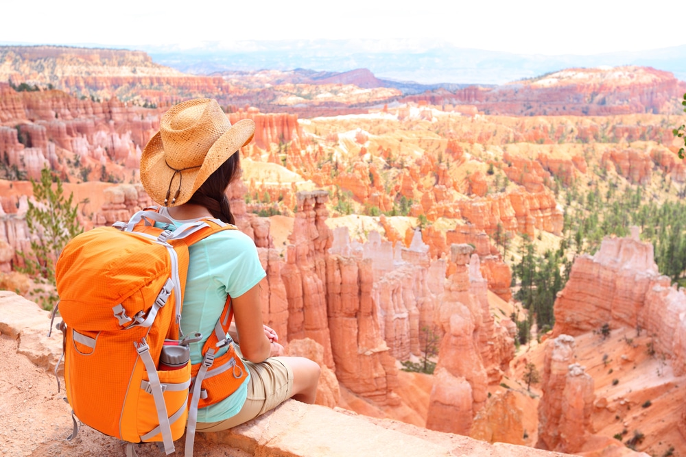 A woman with an orange backpack sits on a cliff overlooking bryce canyon.