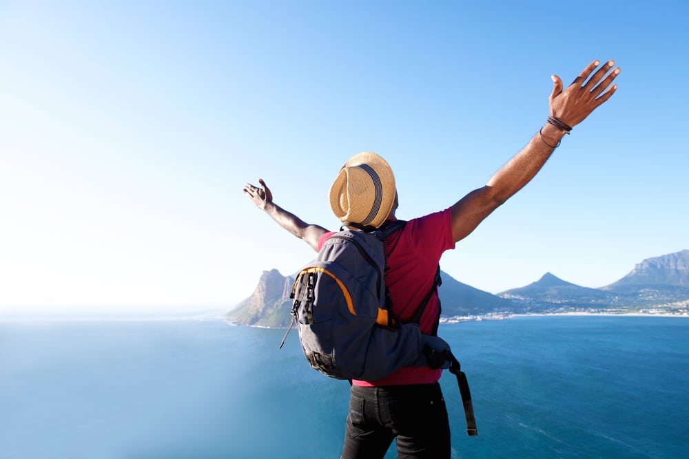 A man with a backpack standing on top of a mountain overlooking the ocean, enjoying the National Thrive Outside Day.
