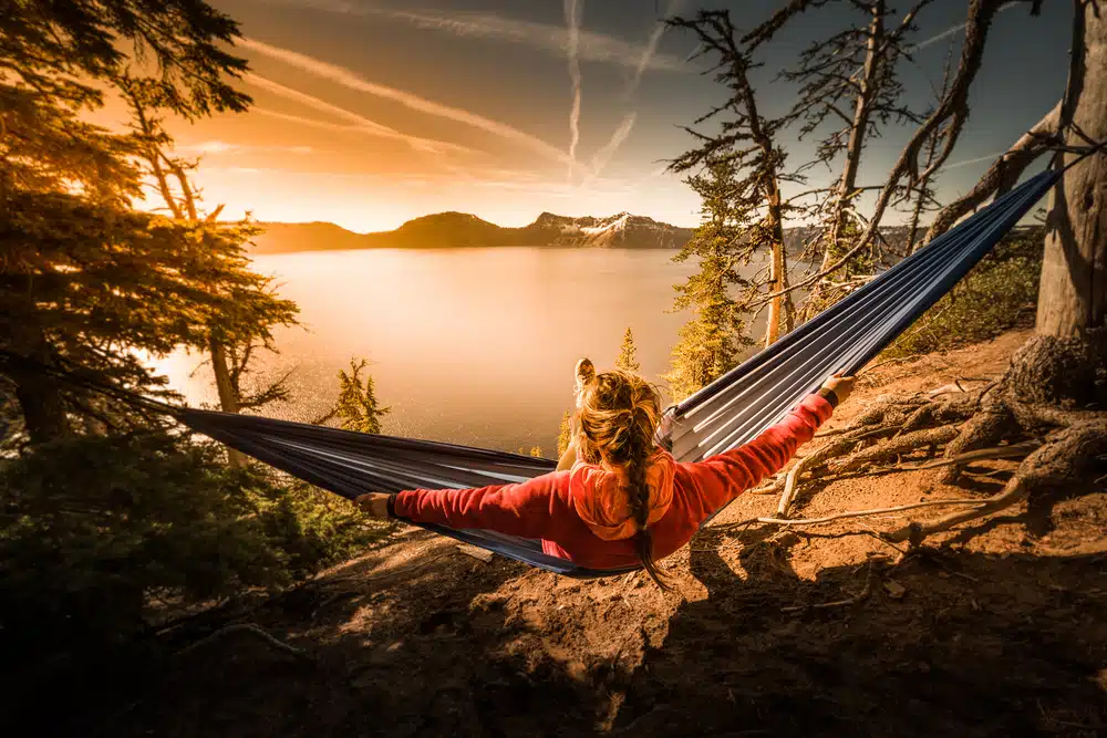 A woman enjoying outdoor recreation relaxes in a hammock overlooking crater lake at sunset.