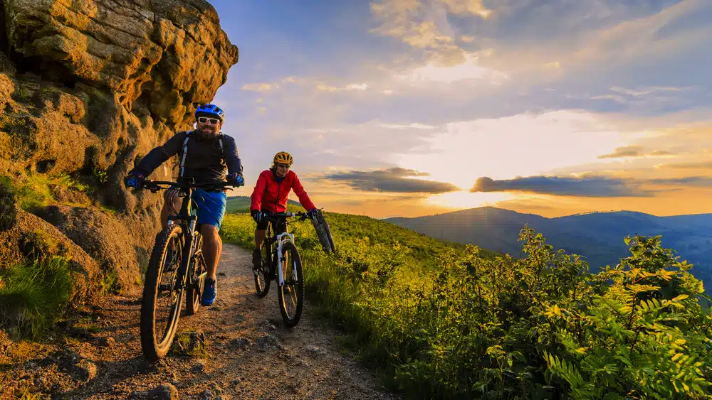 Two people enjoying recreation wins as they ride mountain bikes on a trail at sunset.