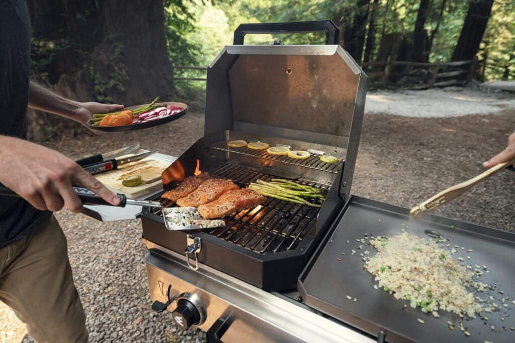 A man is grilling food on an outdoor grill in the woods.