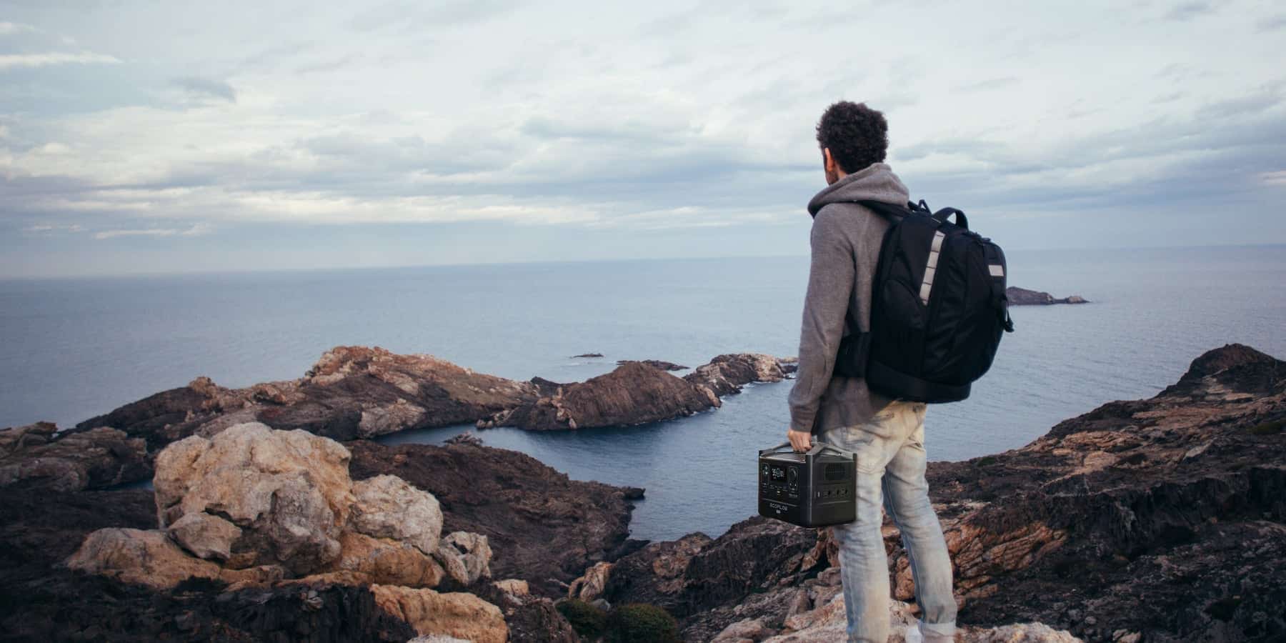 A man with a backpack standing on top of a cliff, using EcoFlow to meet his power needs while gazing at the ocean.