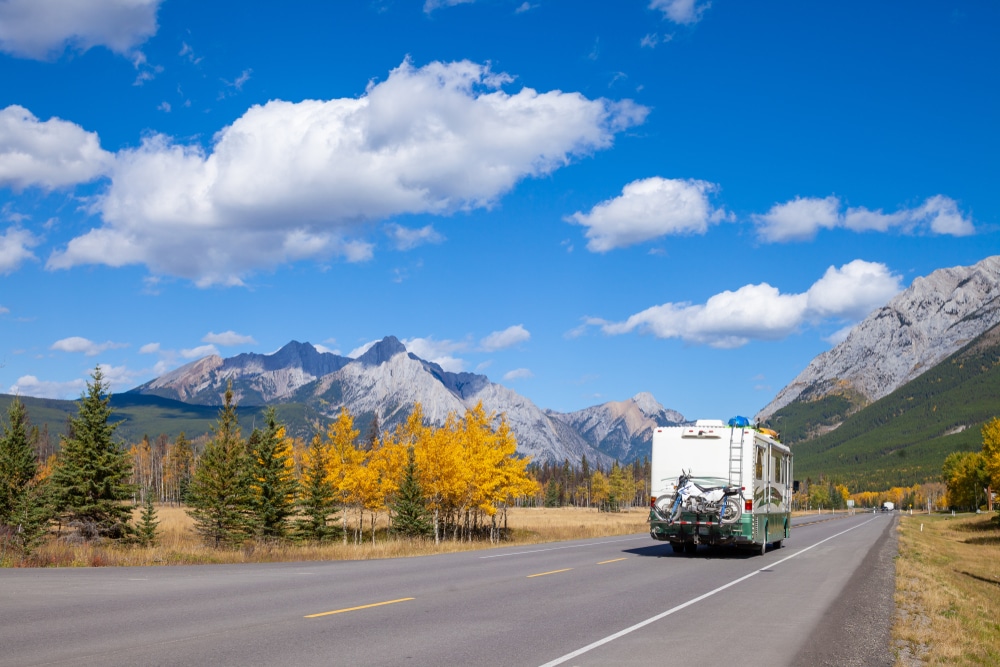 A rv traveling down a road with mountains in the background, showcasing the Canadian Outdoor Hospitality Conference and Expo scene.