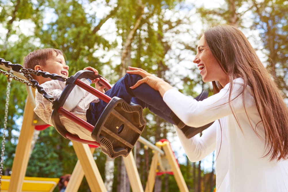 A woman is playing with her son on a swing in a park.