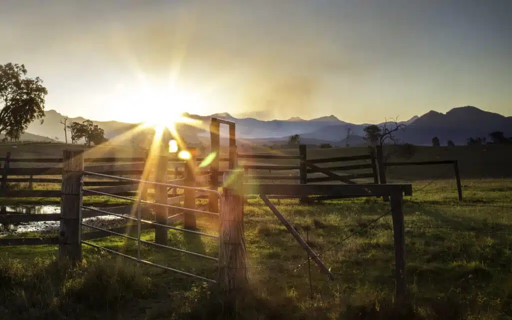 The sun is setting behind a fence in an Australian farm field with mountains in the background.