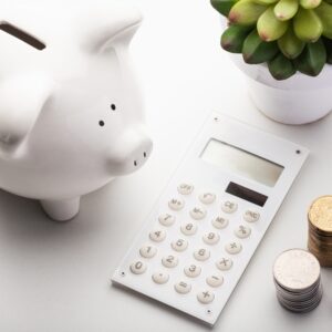A piggy bank filled with coins sits next to a plant on a desk, while a calculator is used for digital financing calculations.