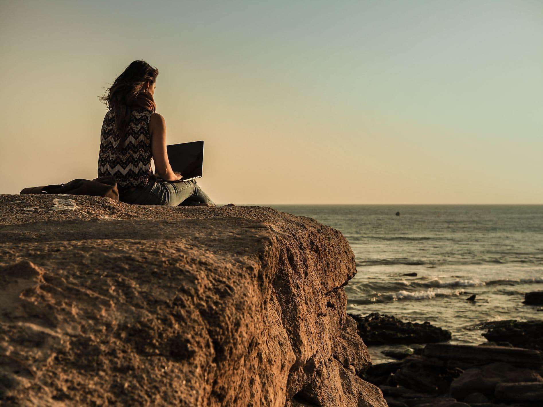 A woman sitting on a cliff looking at the ocean while working on her laptop for a Workcation, finding the cure for Vacation Deprivation.
