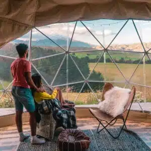 Two people sitting inside a dome tent looking out over a valley.