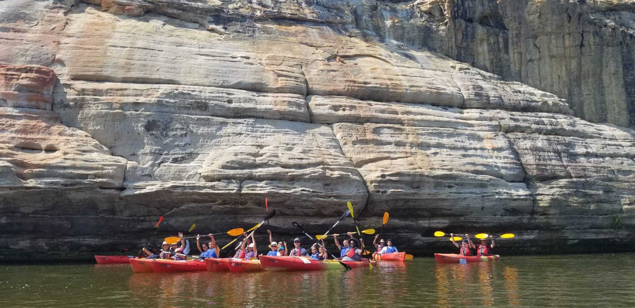 A guided group of people paddling kayaks in front of a cliff at Starved Rock Campground.
