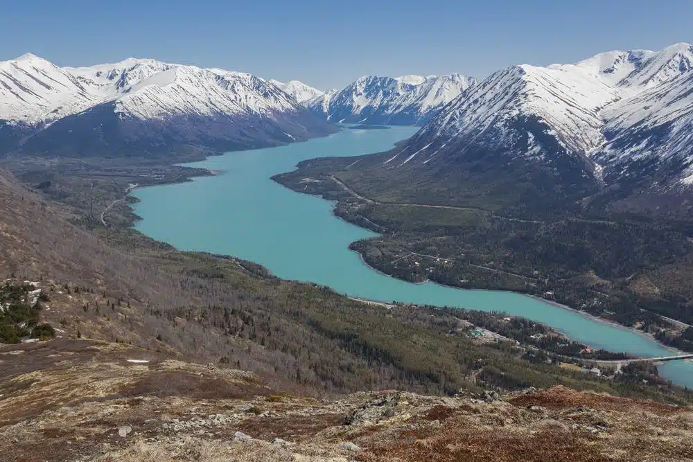 A breathtaking view from the top of a mountain in Alaska, showcasing a serene lake and majestic snow capped mountains.