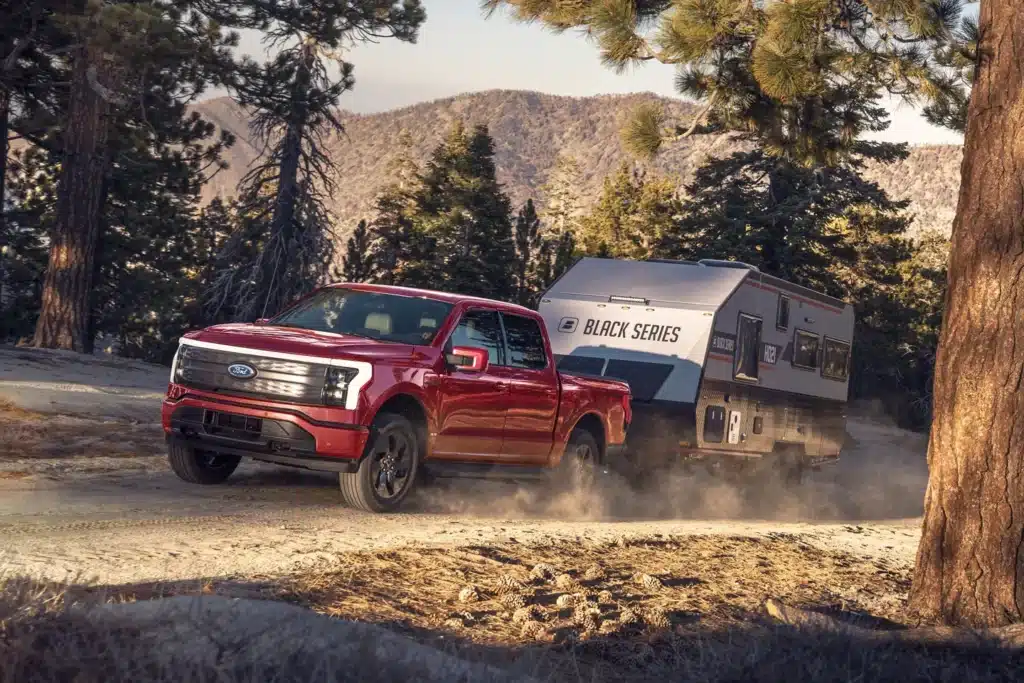 The 2020 Ford F-150 Lightning, an electric vehicle by Ford, is driving down a dirt road with a trailer behind it.
