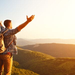 A man standing on top of a mountain in NSW with his arms outstretched, enjoying the breathtaking holiday experience offered by regional tourism.