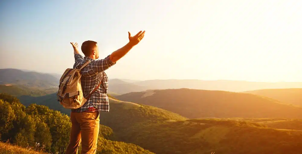 A man standing on top of a mountain in NSW with his arms outstretched, enjoying the breathtaking holiday experience offered by regional tourism.