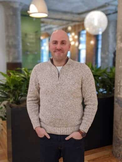 An MC in a sweater standing in an office.