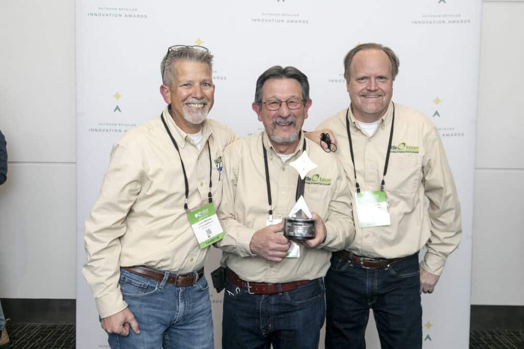 Three men posing for a picture in front of a banner at the awards ceremony for the Little Kamper Program.