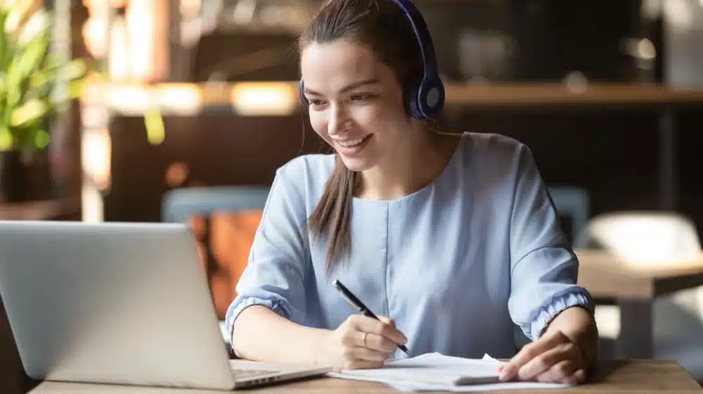 A woman wearing headphones is working on a laptop in a cafe while participating in a Live webinar.