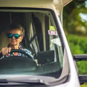 A woman driving an RV with sunglasses on, heading towards Alaska to escape rising gas prices.