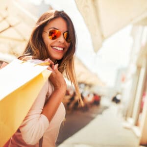 A woman wearing sunglasses and holding shopping bags on a European street, enjoying the hot summer deals.