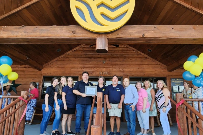 Un grup de oameni care participă la o ceremonie de tăiere a panglicii în fața clădirii Sun Outdoors Chesapeake Bay și își afișează noul nume.