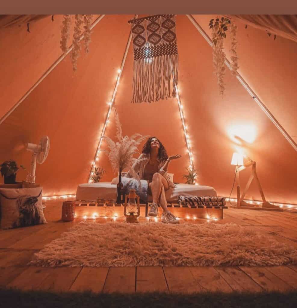 A glamorous woman sitting in a teepee at a glamping site adorned with lights.