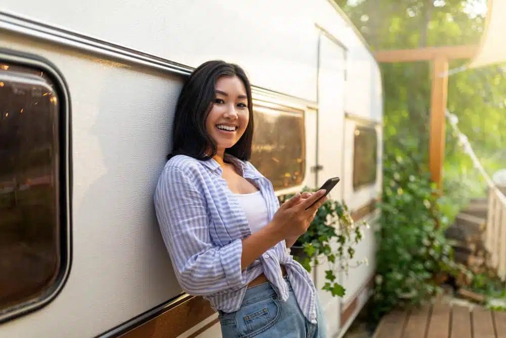 A young woman holding a cell phone in front of a camper trailer while using an Android app.