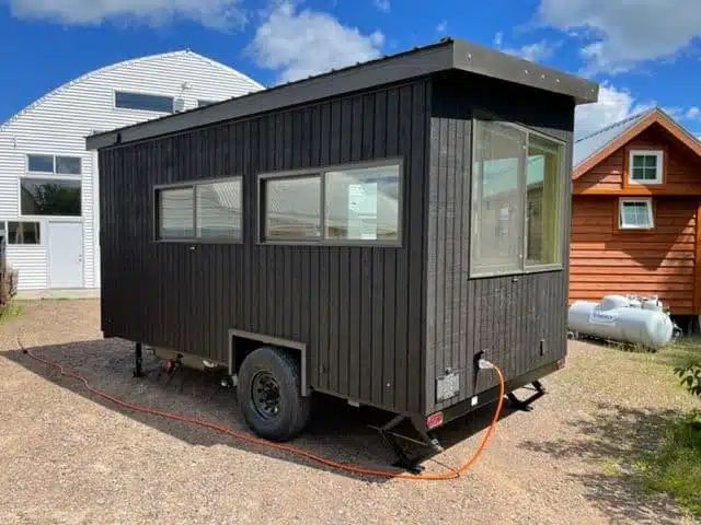 A black ESCAPE Homes tiny house, measuring 120-square foot and entirely all-electric, is parked in front of a house.