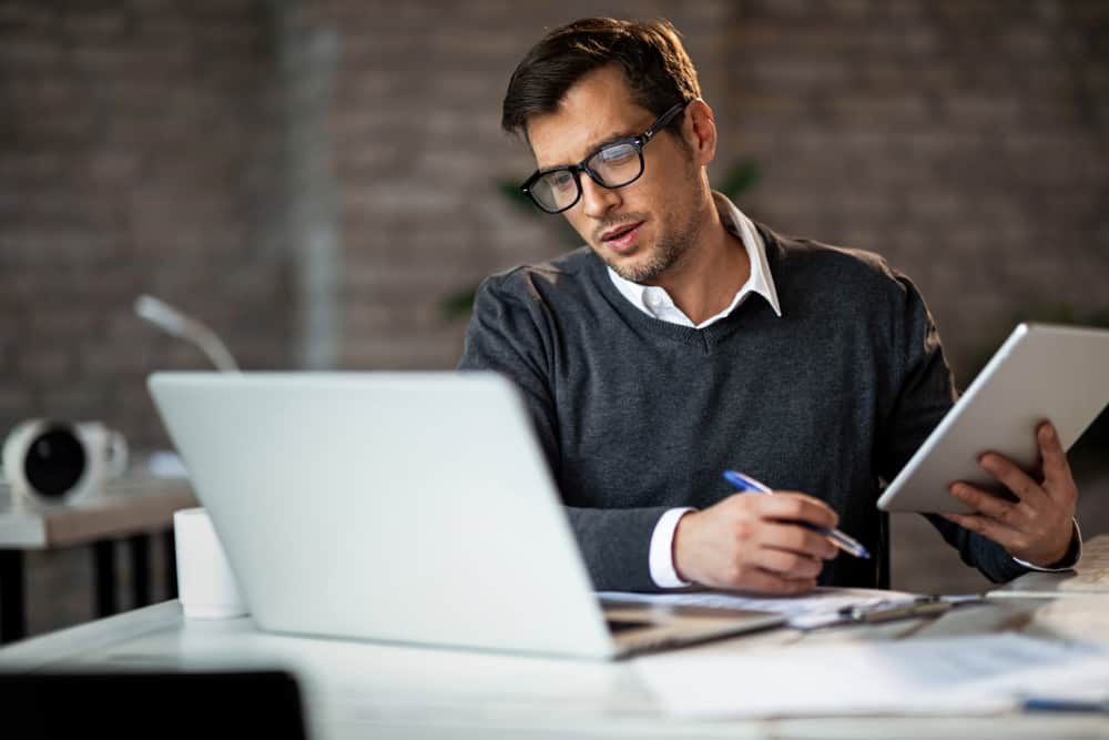 A man in glasses is sitting at a desk with a laptop.