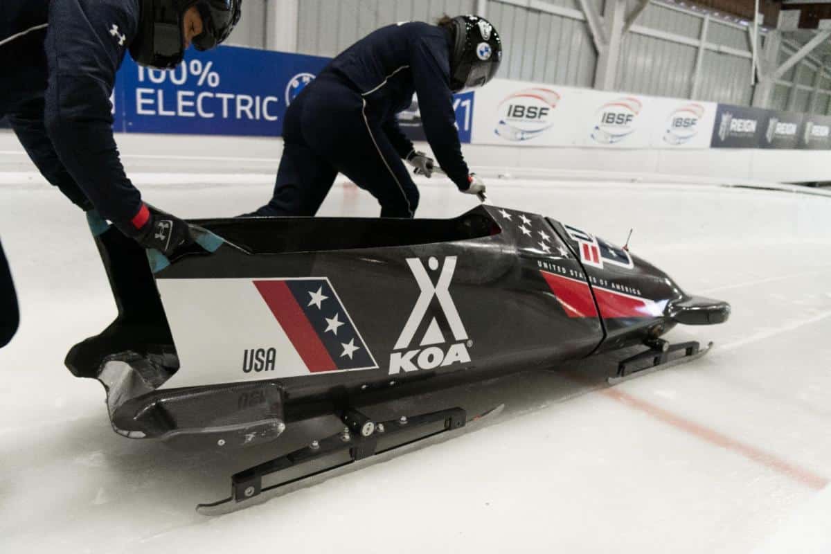 A pair of people standing next to a sled on an ice rink, showcasing their USA Bobsled/Skeleton partnership with KOA sponsorship.