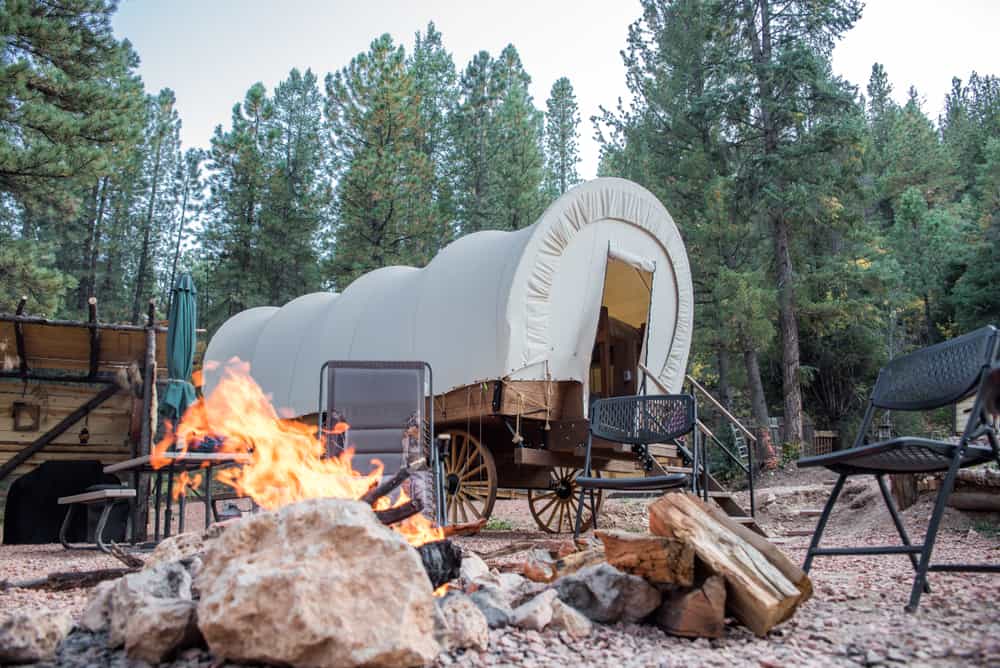 A covered wagon sits next to a fire pit in the woods, creating highlights of glamping ambiance.