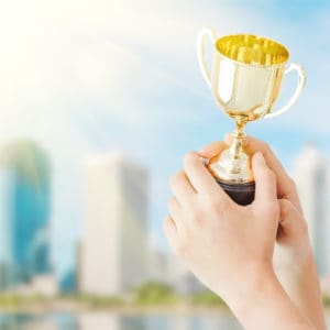 A hand holding a gold trophy representing the 2023 COHCE Award Nominations criteria, set against the backdrop of a city.