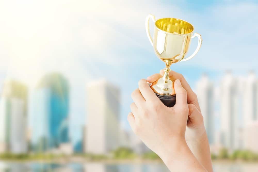 A hand holding a gold trophy representing the 2023 COHCE Award Nominations criteria, set against the backdrop of a city.