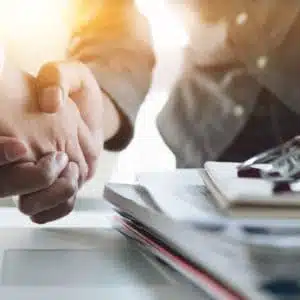 Two business people shaking hands in front of a laptop, finalizing the acquisition deal.