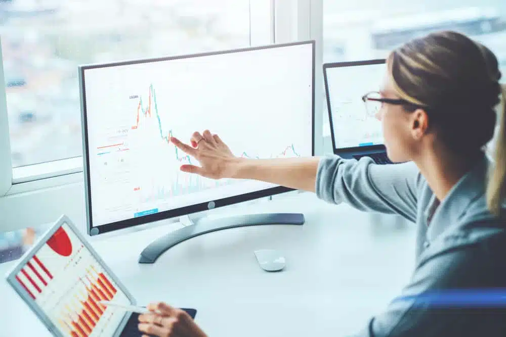 A Financial Analyst from IVEE Group is pointing at a graph on a computer screen in a team meeting.
