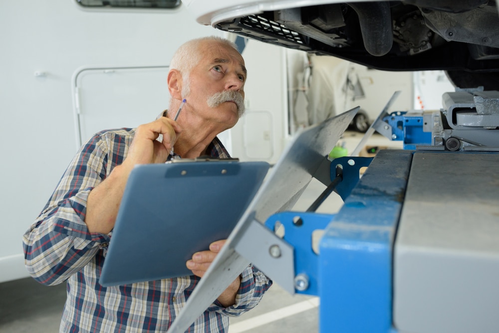 An older man, participating in KOA's Roadside Assistance Program, diligently inspects a clipboard under the hood of a vehicle.