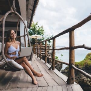 A woman enjoying experiential travel on a swing chair with a laptop on the beach.
