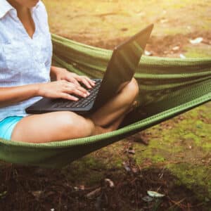 A woman is sitting in a hammock using her laptop while enjoying the peaceful surroundings provided by Good Sam.