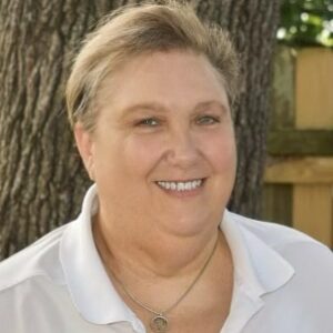 A woman in a white shirt smiling in front of a tree during MC Fireside Chats on August 16th.