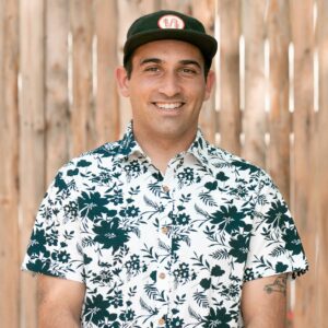 A man in a floral shirt standing in front of a wooden fence on February 28th, 2024.