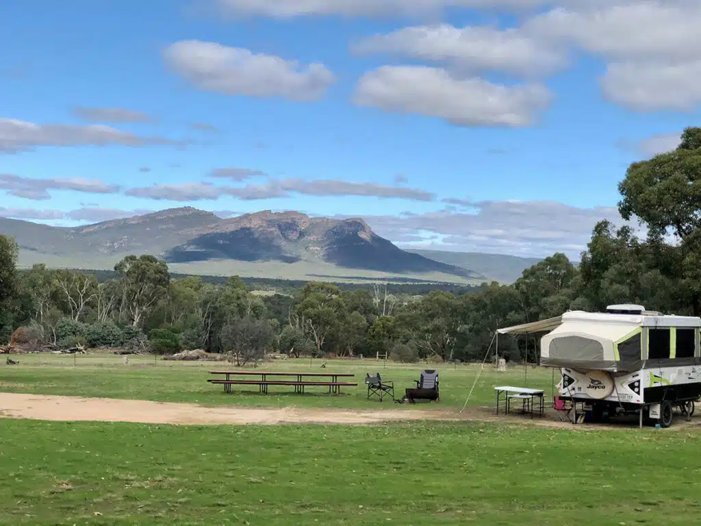 An RV parked in a grassy area with mountains and log cabins in the background.