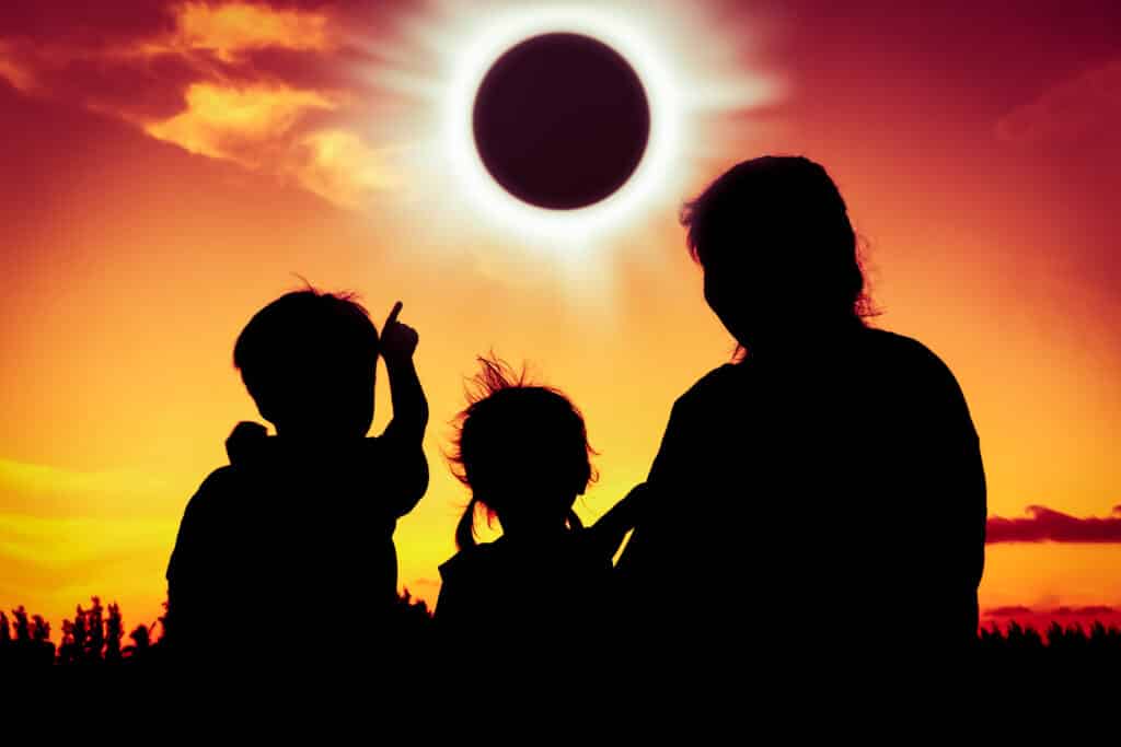 Silhouetted family at a campspot watching a solar eclipse at sunset, with one child pointing upwards at the darkened sun.