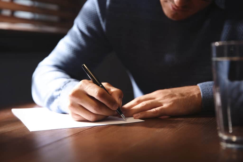 A man in a blue sweater writing on a white sheet of paper at a wooden table, with a glass of water beside him, focusing on rural economies.