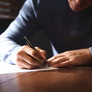 A man in a blue sweater writing on a white sheet of paper at a wooden table, with a glass of water beside him, focusing on rural economies.